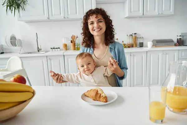 Mujer alegre cogida de la mano de un niño lindo cerca de croissant y jugo de naranja con frutas en la cocina - foto de stock
