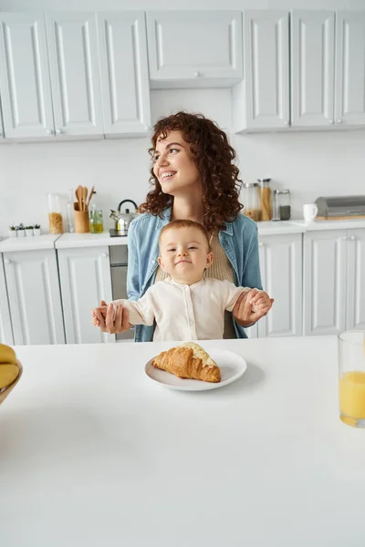 Smiling woman looking away and holding hands of happy child near croissant and fresh orange juice — Stock Photo
