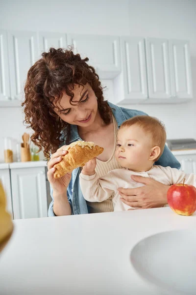 Curly woman giving delicious croissant to toddler daughter near fresh apple, breakfast in kitchen — Stock Photo