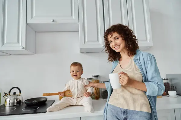 Женщина с чашкой кофе улыбается перед камерой рядом с малышкой, сидящей на кухонном столе с деревянной вилкой — стоковое фото