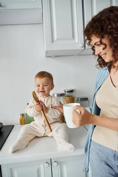 Enfant gai assis sur le plan de travail de la cuisine et jouer avec une fourchette en bois près de maman heureuse avec tasse de café — Photo de stock