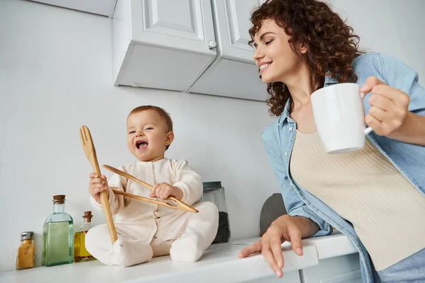 Donna sorridente con tazza di caffè vicino bambino felice giocando con posate in legno sul piano di lavoro della cucina — Foto stock