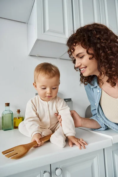 Cheerful woman near little daughter with wooden fork sitting on kitchen worktop, happy morning — Stock Photo