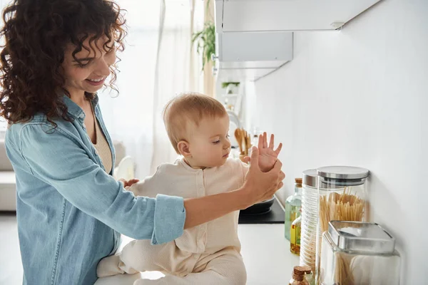 Mujer feliz sosteniendo la pequeña mano de la hija del niño sentado en el mostrador de la cocina, la paternidad dichosa - foto de stock