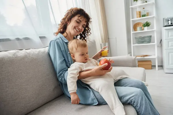 Mujer alegre sosteniendo manzana y jugo de naranja mientras está sentado en un sofá acogedor con su hija pequeña - foto de stock