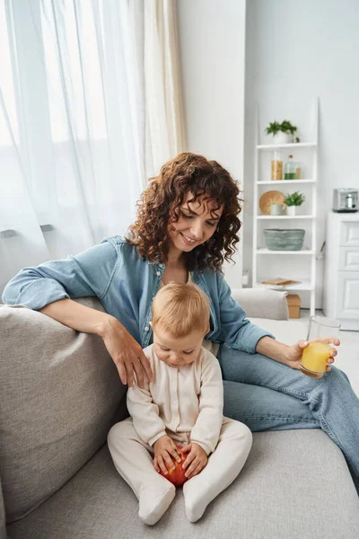Smiling woman with glass of orange juice sitting on couch near cute baby girl with fresh apple — Stock Photo