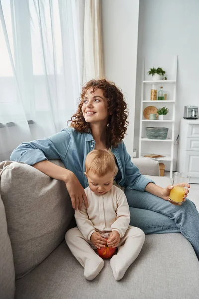 Enchantée femme avec du jus d'orange regardant loin sur le canapé près adorable enfant avec pomme, bonheur — Photo de stock