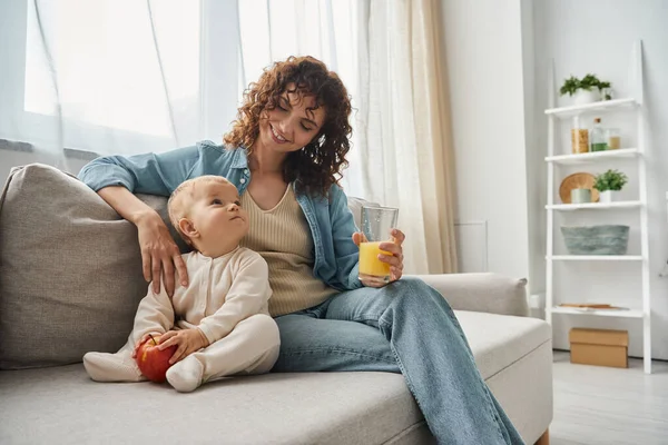 Mother and toddler girl with orange juice and fresh apple looking at each other on cozy couch — Stock Photo