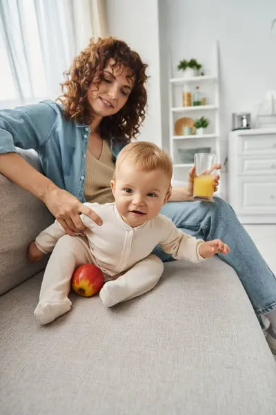 Aufgeregtes kleines Mädchen blickt in die Kamera neben Apfel und Mutter mit Orangensaft auf der heimischen Couch — Stockfoto