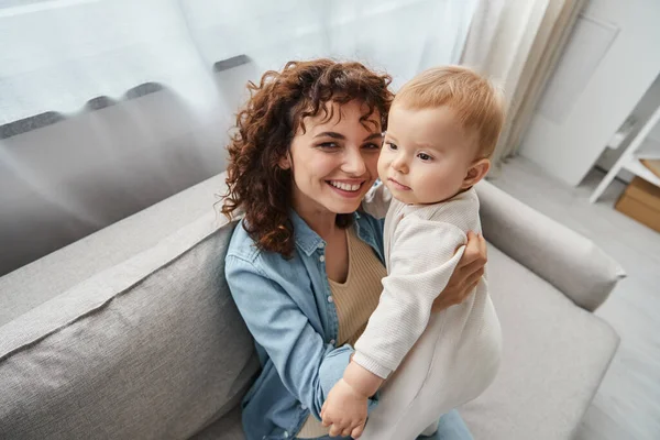 Femme joyeuse tenant adorable petite fille et regardant la caméra sur le canapé à la maison, parentalité heureuse — Photo de stock