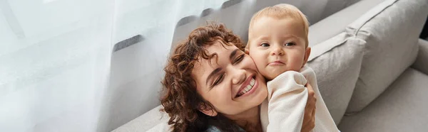 Happy mother with closed eyes holding adorable toddler child on couch in living room, banner — Stock Photo