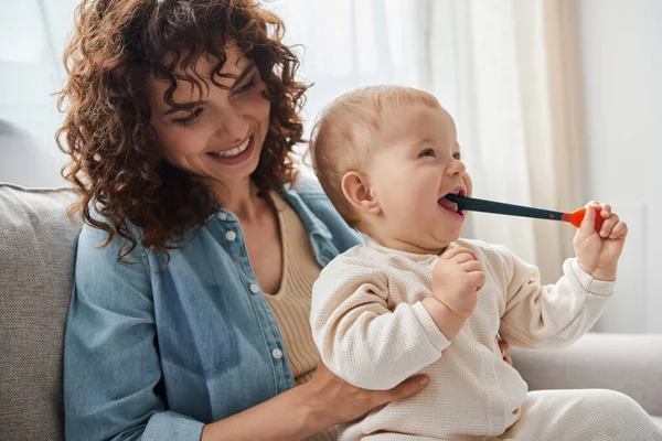 Lächelnde Frau auf Couch sitzend mit Kleinkind-Tochter, die Kinderspielzeug im Wohnzimmer kaut, Spielzeit — Stockfoto