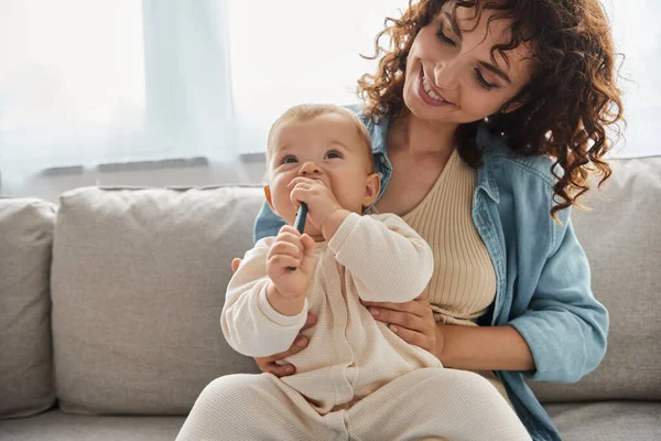 Adorable baby chewing teething toy near cheerful mother on couch in living room, leisure and fun — Stock Photo
