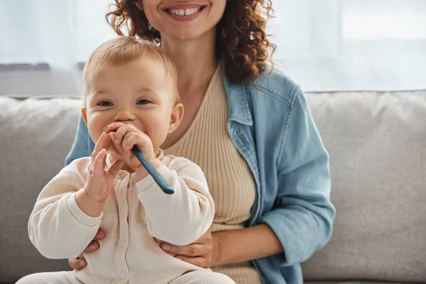 Bambino spensierato masticare giocattolo dentizione vicino sorridente madre sul divano in soggiorno, infanzia beata — Foto stock