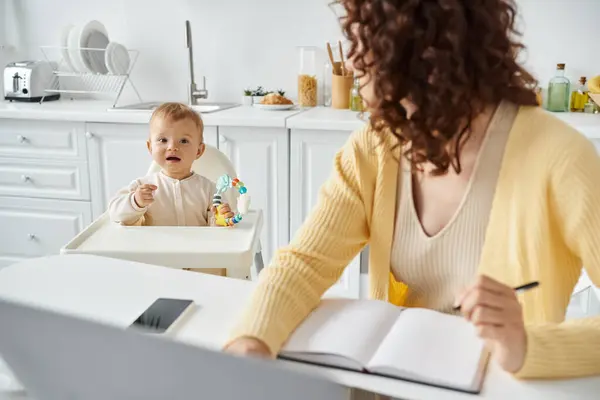 Girl with rattle toy looking at mom working near laptop and smartphone in kitchen, work life balance — Stock Photo