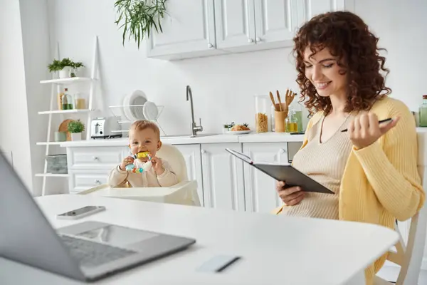 Happy woman sitting with notebook near laptop and toddler kid with rattle toy in cozy kitchen — Stock Photo