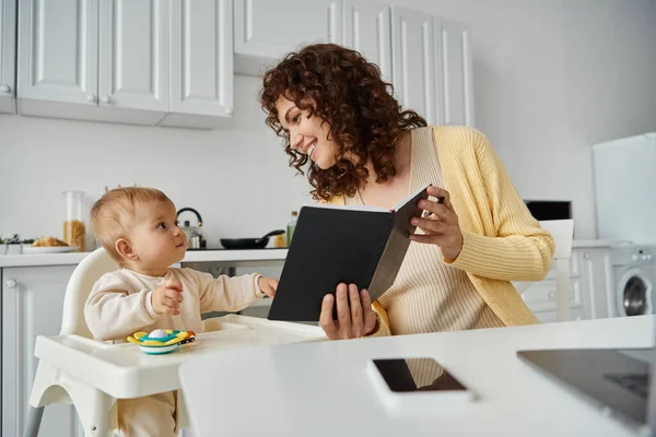 Cheerful woman showing notebook to little daughter sitting in baby chair in kitchen, working parent — Stock Photo
