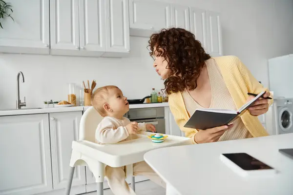 Femme avec ordinateur portable envoyant baiser d'air à la petite fille assise sur une chaise bébé, travaillant dans la cuisine — Photo de stock