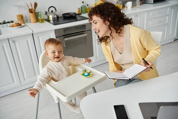 Mãe alegre com notebook perto de criança animada sentada em cadeira de bebê com brinquedo chocalho, pai trabalhando — Fotografia de Stock