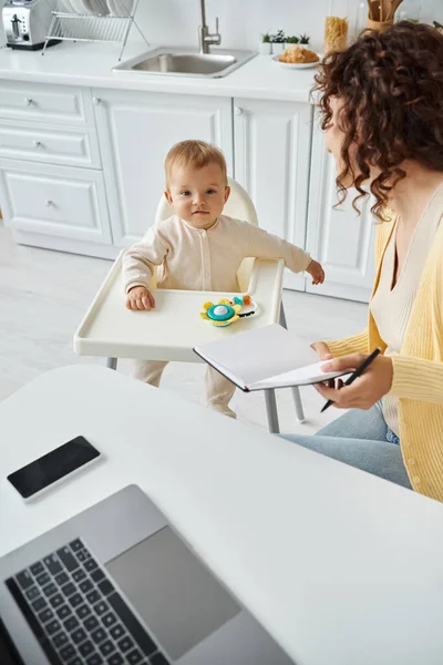 Cute child in baby chair near mother working with notebook and laptop in kitchen, work life balance — Stock Photo
