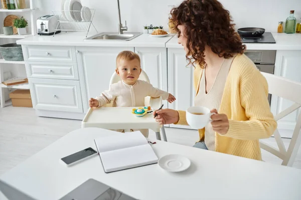 Frau mit Kaffeetasse sitzt neben Notizbuch und Smartphone neben Kleinkind-Tochter im Babystuhl — Stockfoto