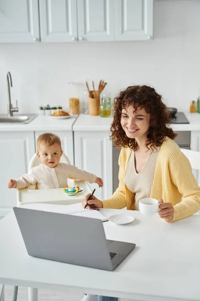Mulher positiva com xícara de café olhando para laptop enquanto trabalhava na cozinha perto da filha da criança — Fotografia de Stock