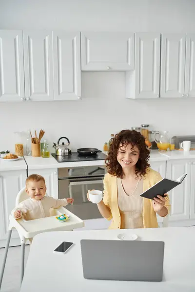 Mulher positiva com xícara de café e notebook olhando para laptop perto de criança despreocupada na cozinha — Fotografia de Stock