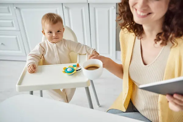 Mujer con café de la mañana y portátil cerca de niño sonriente en silla de bebé con juguete sonajero en la cocina - foto de stock