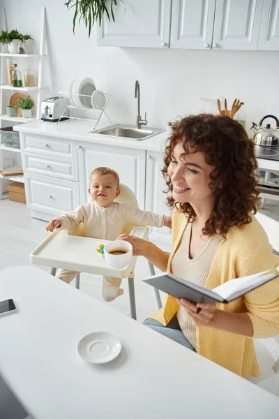 Smiling woman with morning coffee and notebook near cheerful child sitting in baby chair in kitchen — Stock Photo