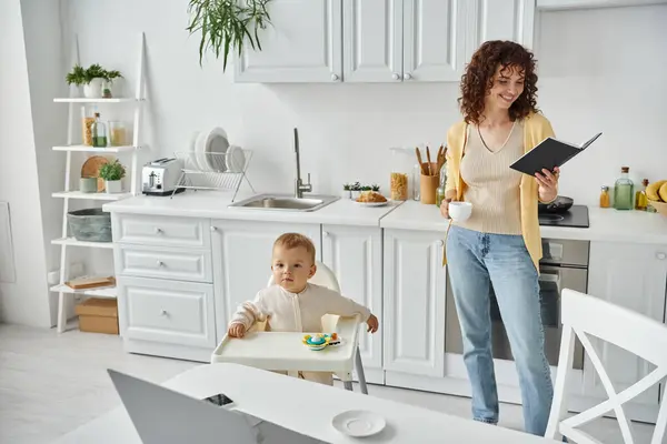Femme joyeuse avec tasse de café à la recherche dans le portable près de l'ordinateur portable et petit enfant dans la chaise de bébé dans la cuisine — Photo de stock