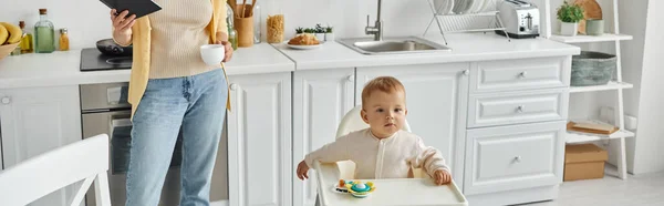 Little girl sitting in baby chair near rattle toy near mom with notebook and coffee cup, banner — Stock Photo