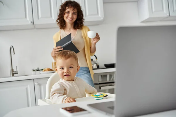Cute baby smiling at camera near mom with coffee cup and notebook and laptop on blurred foreground — Stock Photo