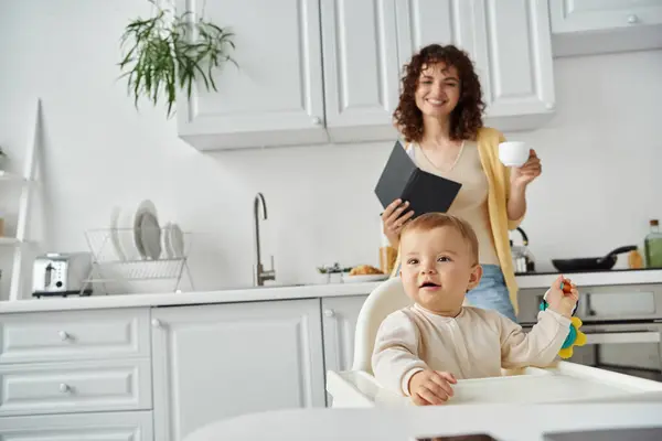 Bambino bambino che gioca con sonaglio giocattolo vicino alla madre con notebook e tazza di caffè in cucina moderna — Foto stock