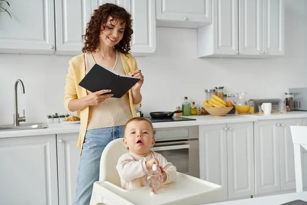 Sorrindo freelancer feminino escrevendo no caderno enquanto a filha da criança brincando com o brinquedo chocalho em casa — Fotografia de Stock