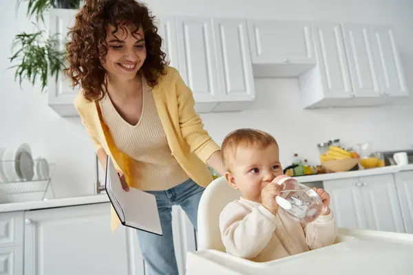 Mãe alegre com notebook olhando para a filha da criança bebendo de mamadeira na cozinha — Fotografia de Stock