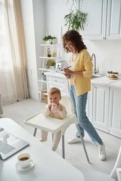 Mujer sonriente escribiendo en el cuaderno mientras que la pequeña hija bebiendo agua del biberón cerca del ordenador portátil - foto de stock