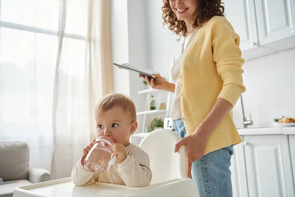 Little girl sitting in baby chair and drinking water near smiling mother with notebook in kitchen — Stock Photo