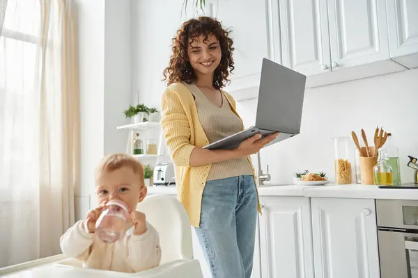 Happy woman with laptop looking at toddler daughter drinking water from baby bottle in kitchen — Stock Photo