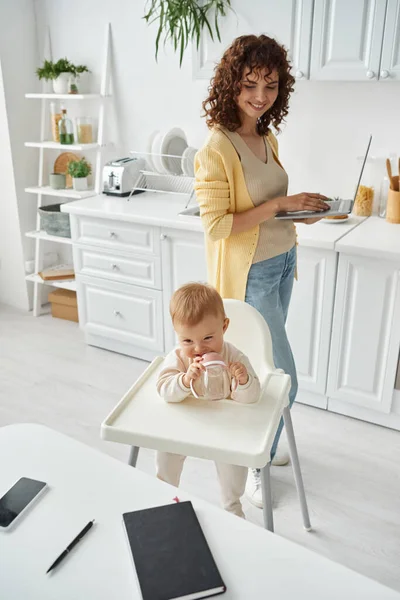 Cute child chewing baby bottle near mother with laptop and notebook with smartphone on kitchen table — Stock Photo