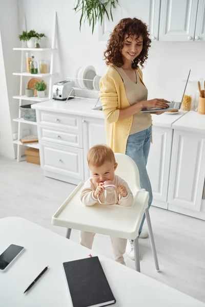 Bonito criança mastigar bebê garrafa perto de mãe com laptop e notebook com smartphone na mesa da cozinha — Fotografia de Stock