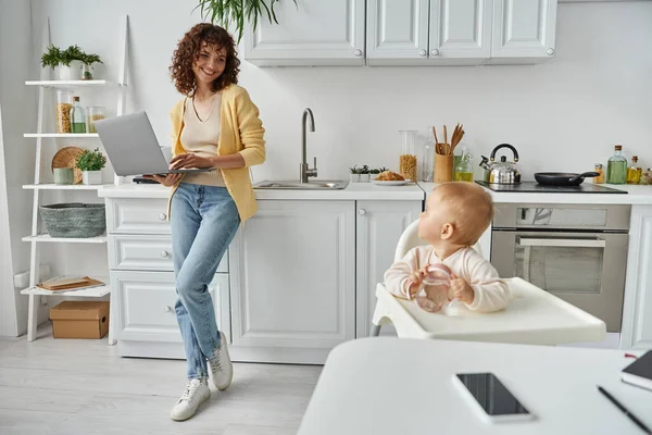 Mère avec ordinateur portable regardant bébé fille assise avec biberon dans la cuisine, travail à la maison — Photo de stock