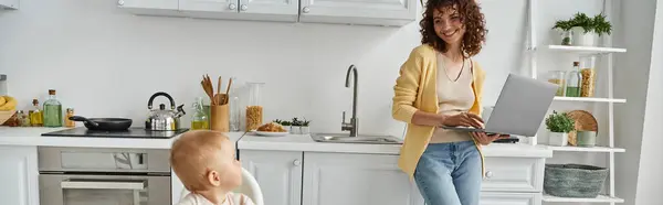 Kleinkind schaut fröhliche Mutter an, die mit Laptop in Küche steht, Work-Life-Balance, Banner — Stockfoto