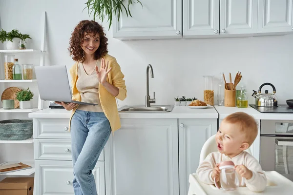 Joyful woman with laptop waving hand to little daughter with baby bottle in kitchen, freelance work — Stock Photo