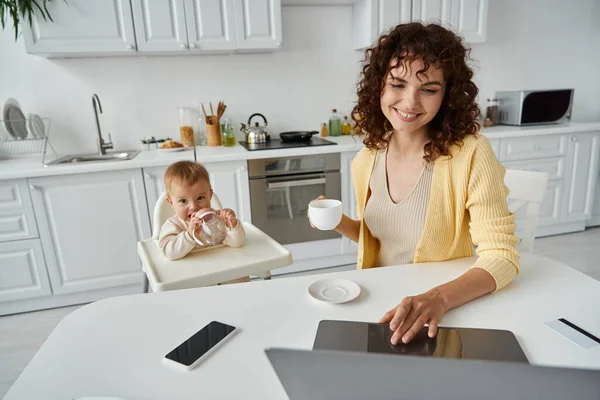 Smiling woman with coffee cup working on laptop near smartphone and little kid with baby bottle — Stock Photo