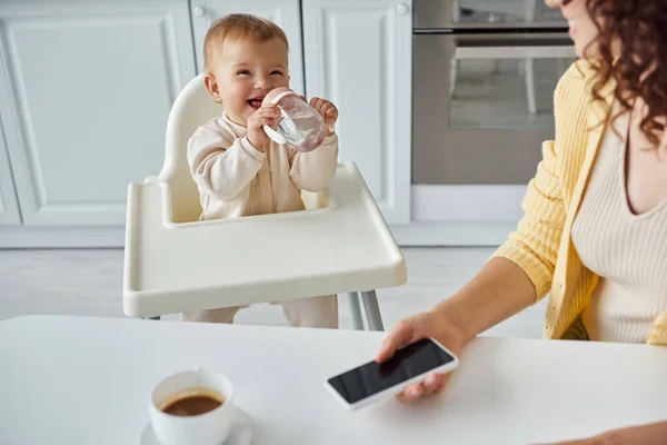 Cheerful child drinking from baby bottle near mom with smartphone with blank screen near coffee cup — Stock Photo