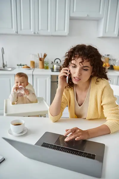 Busy woman talking on smartphone and working on laptop near little child in kitchen, freelance work — Stock Photo