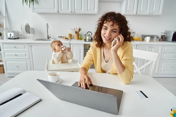 Frau spricht mit Smartphone und arbeitet am Laptop neben Kleinkind in Küche, ausgewogener Lebensstil — Stockfoto