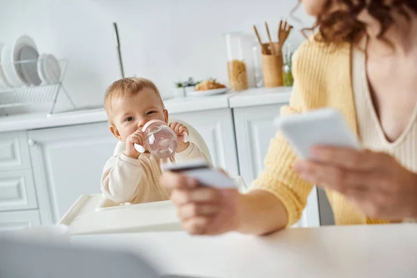 Menina bebendo de mamadeira perto da mãe com smartphone e cartão de crédito em primeiro plano turvo — Fotografia de Stock