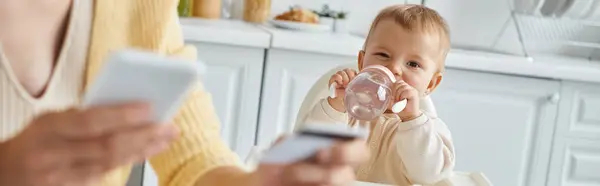 Baby girl drinking from baby bottle near blurred mom with smartphone and credit card, banner — Stock Photo