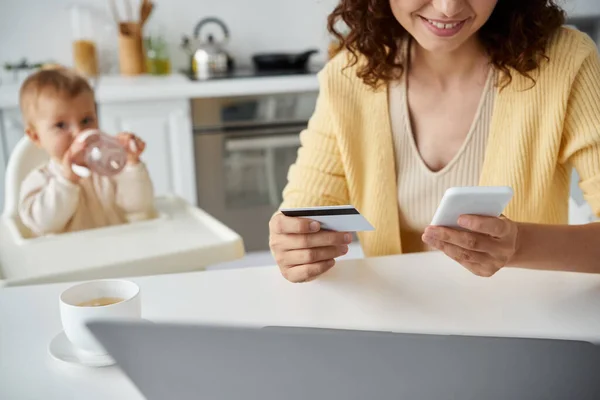 Mulher sorridente com cartão de crédito e smartphone perto da pequena filha beber de mamadeira na cozinha — Fotografia de Stock